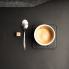 Sous tasse en cuir noir avec tasse de café, cuillère et sucre posés sur un bureau élégant.
