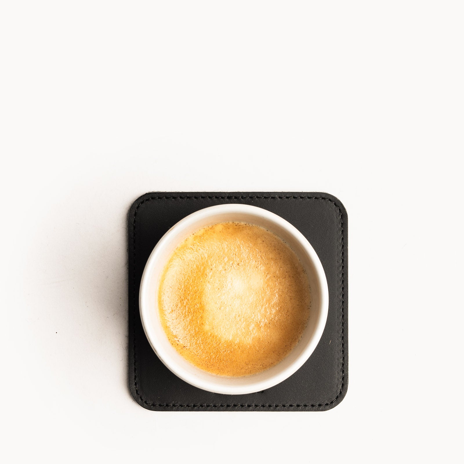 Dessous de verre noir en cuir avec tasse de café vue du dessus, posé sur fond blanc minimaliste.